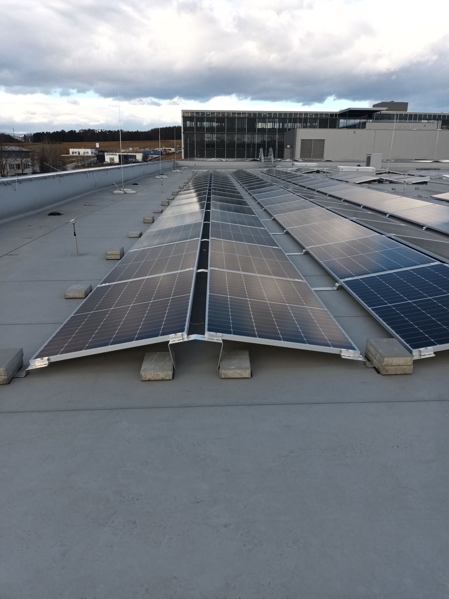 Rząd paneli słonecznych zamontowanych na płaskim dachu budynku przemysłowego, zabezpieczonych betonowymi blokami, z widokiem na sąsiedni budynek biurowy i pochmurne niebo.