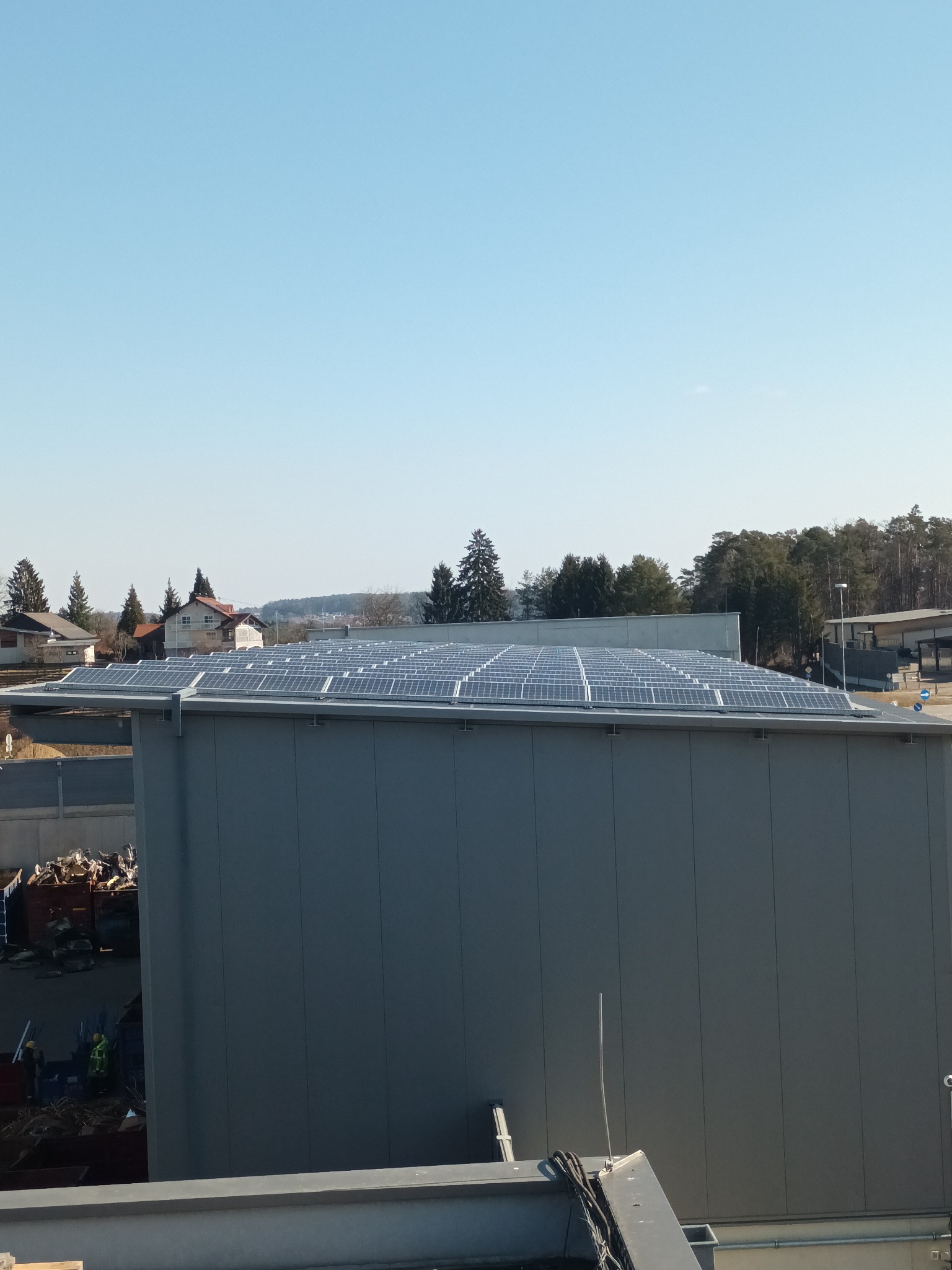 Instalacja paneli słonecznych na dachu szarego budynku przemysłowego, widok z góry, w tle zabudowania mieszkalne i drzewa.