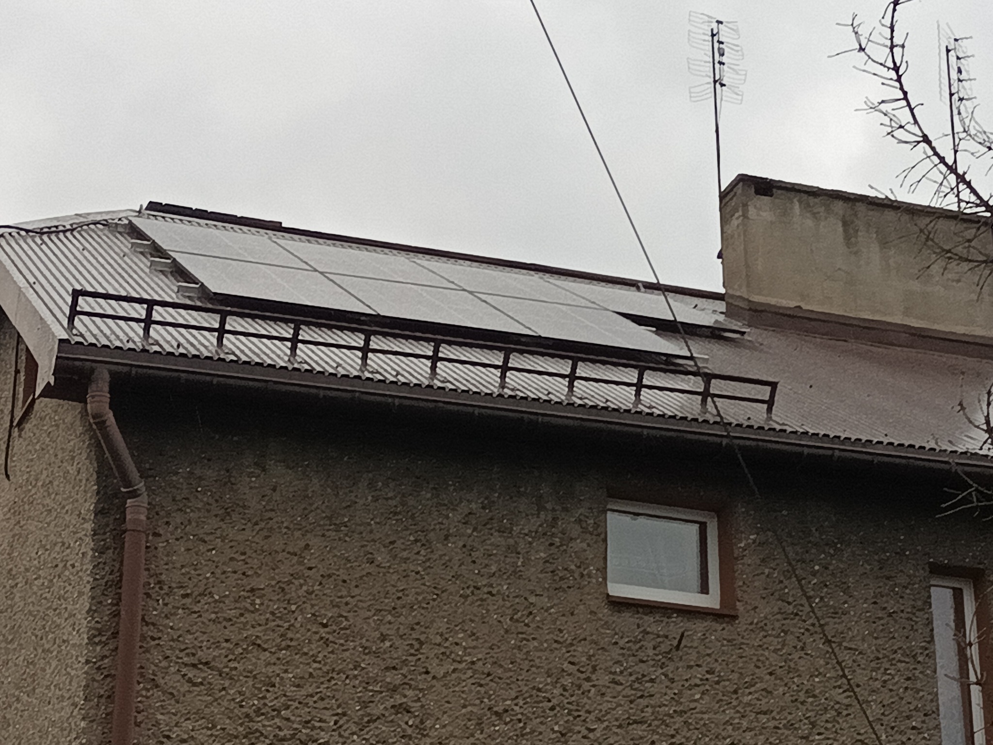 Panele słoneczne zamontowane na metalowym dachu budynku mieszkalnego z anteną telewizyjną w tle, pochmurny dzień.