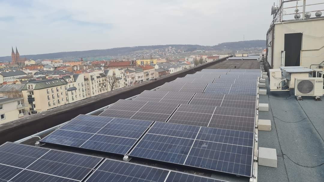 Rząd paneli fotowoltaicznych na płaskim dachu budynku z widokiem na miasto i kościelne wieże w tle, obok jednostki klimatyzacyjne marki Kaisai.