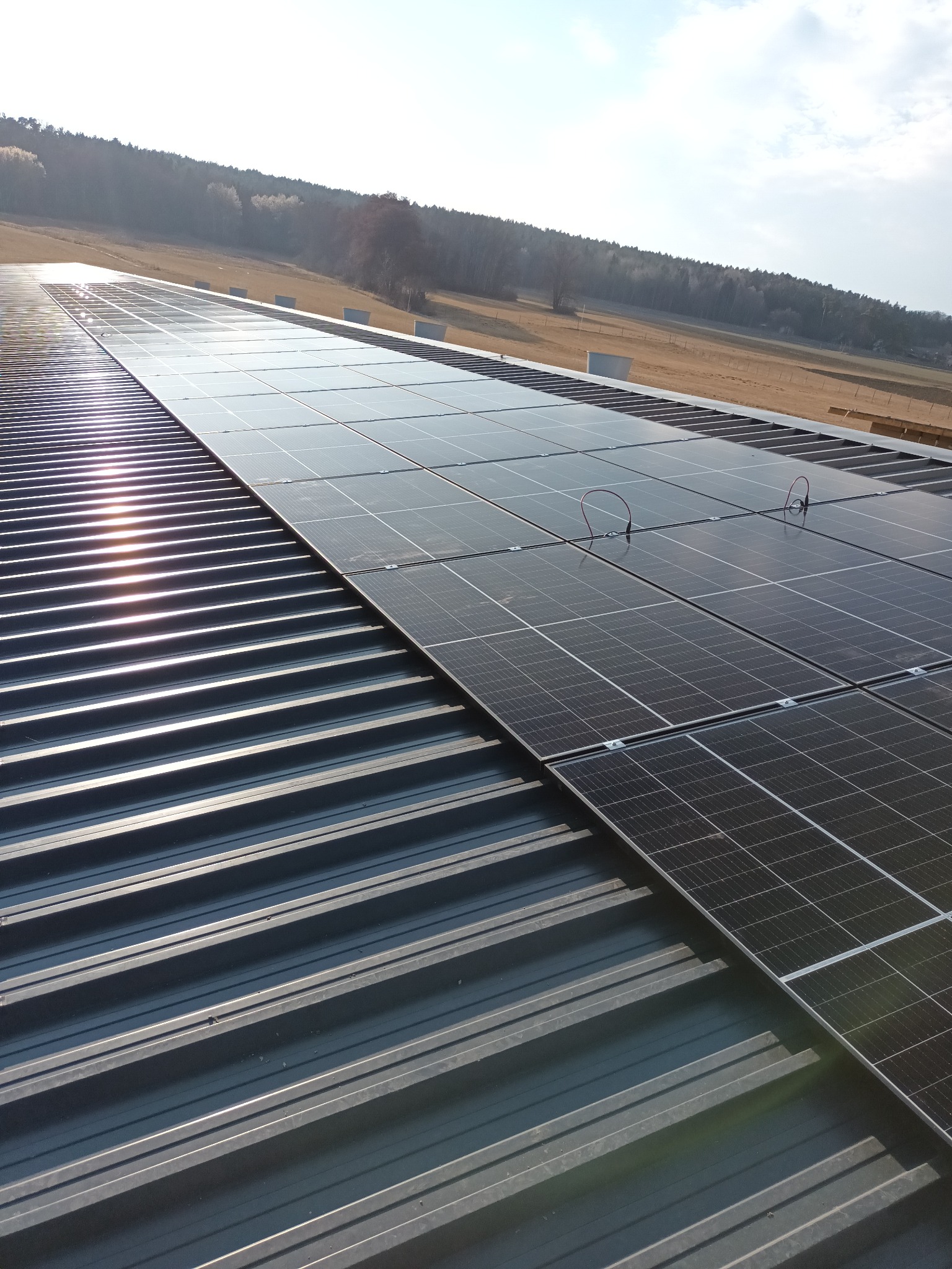 Panel słoneczny na metalowym dachu trapezowym, z widocznymi przewodami i odbiciem słońca, w tle krajobraz z polami i lasem.