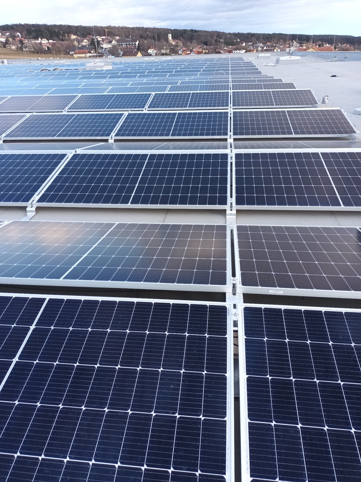 Rozległa instalacja paneli słonecznych na płaskim dachu, z widokiem na odległe zabudowania miejskie pod pochmurnym niebem.