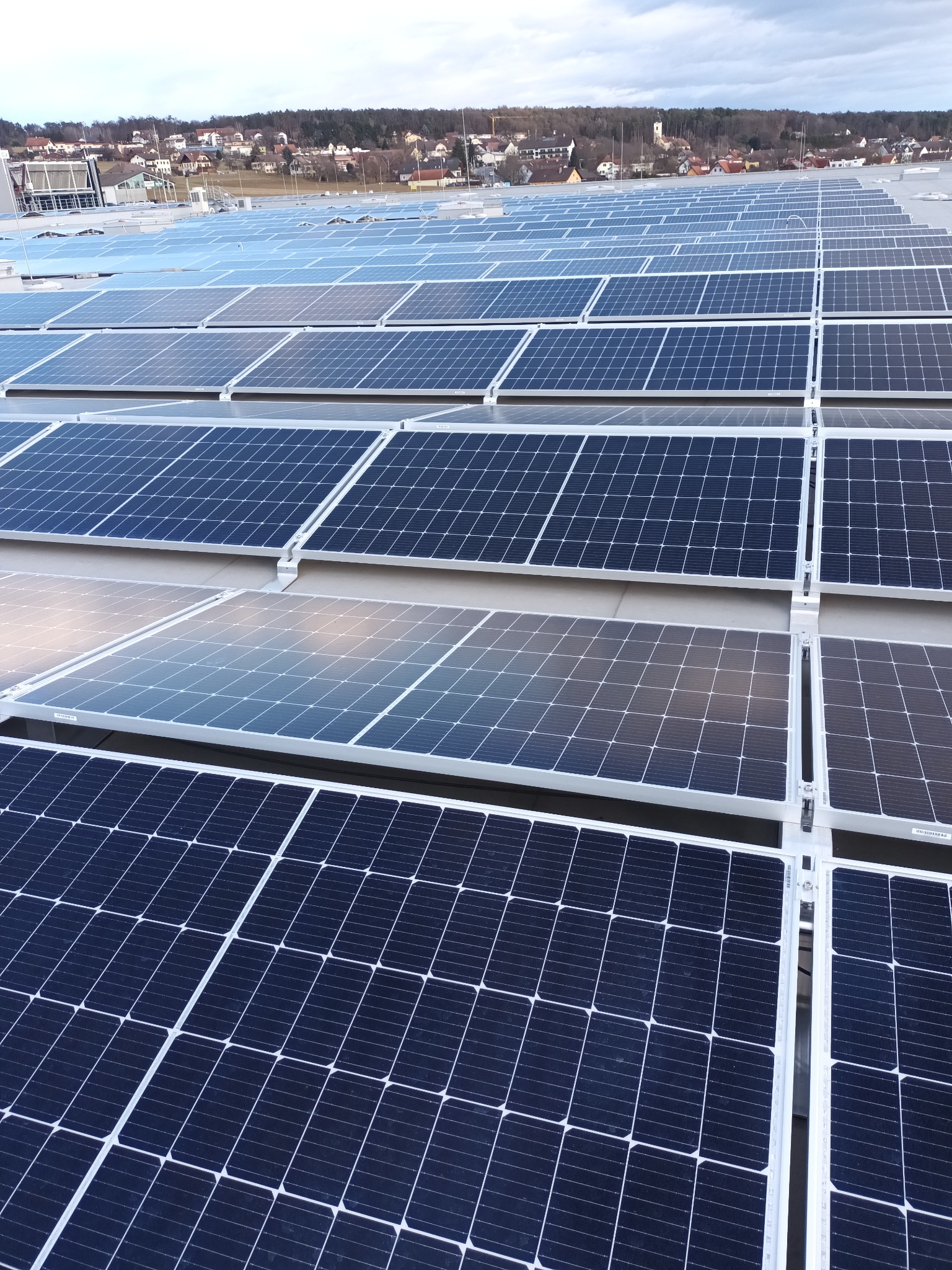 Rozległa instalacja paneli słonecznych na dachu budynku, z widokiem na oddalony krajobraz miejski pod pochmurnym niebem.