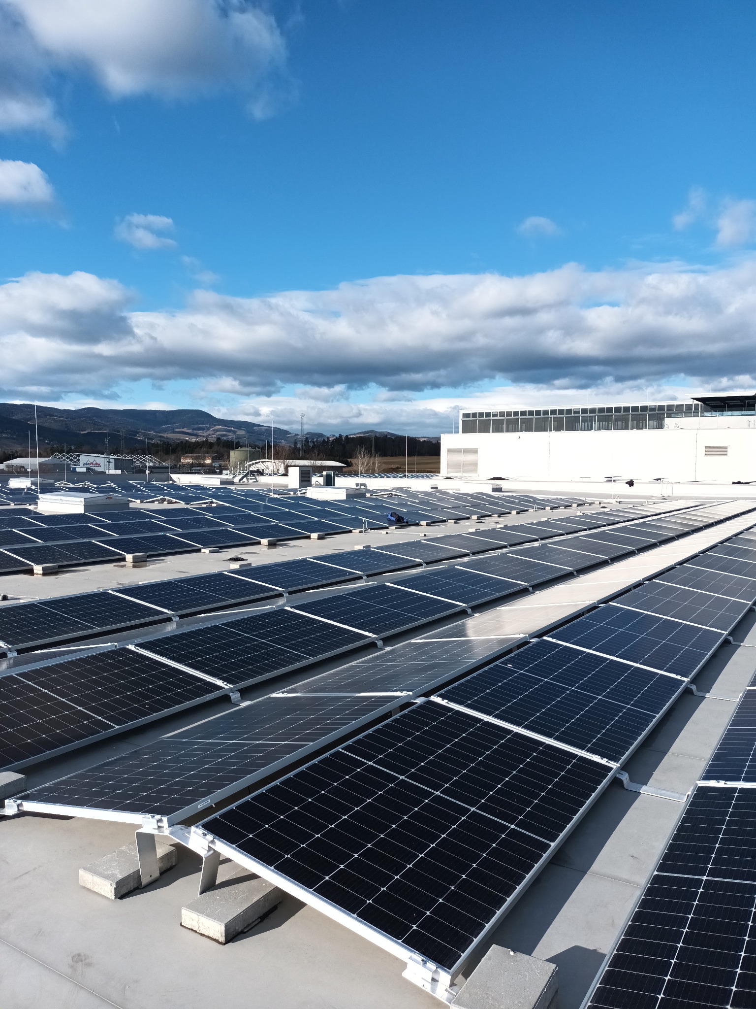 Rząd paneli słonecznych na płaskim dachu budynku przemysłowego, widok z góry w słoneczny dzień z chmurami.