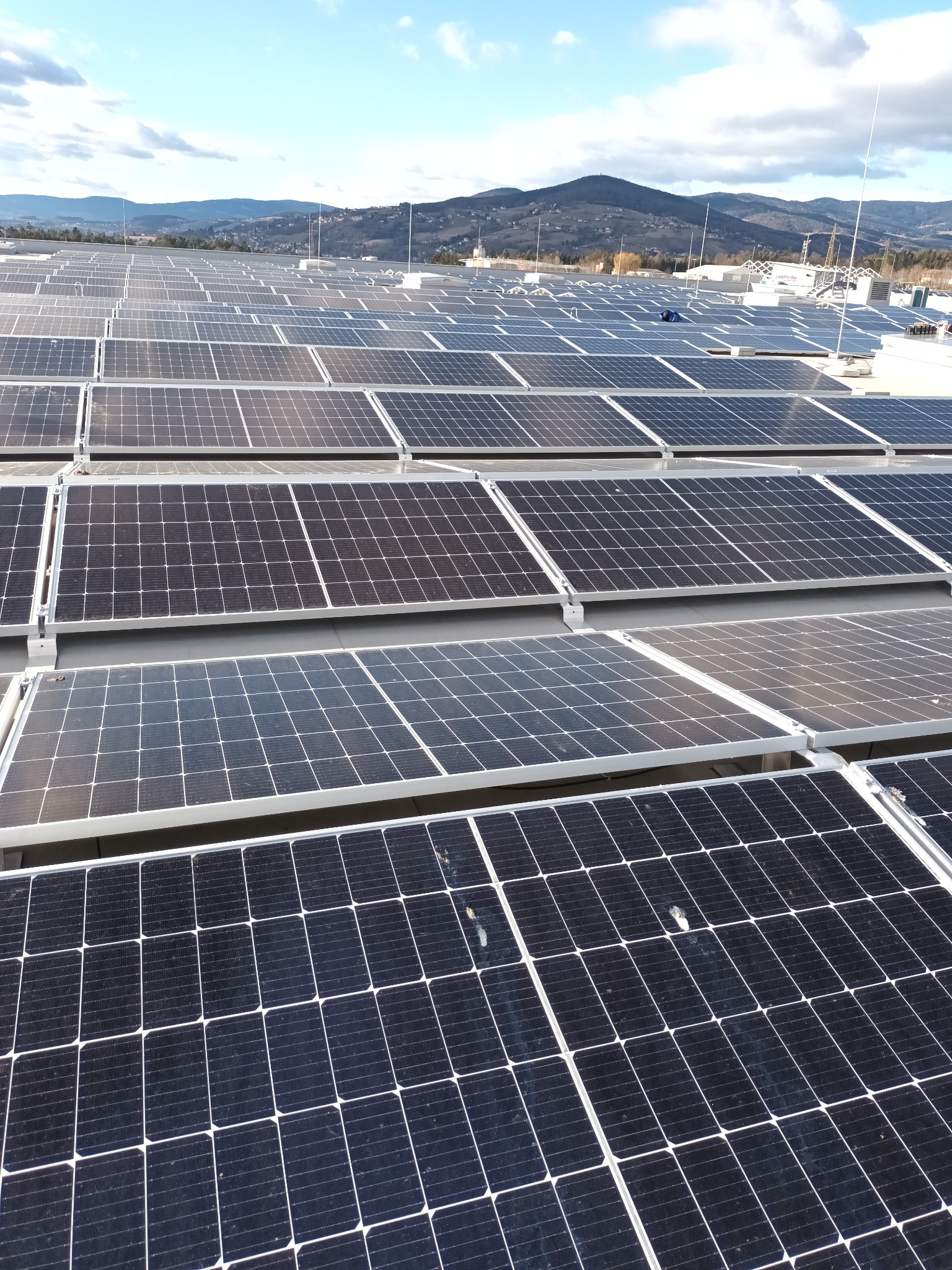Rozległa instalacja paneli słonecznych na dachu budynku, z widocznymi antenami i górami w tle pod błękitnym niebem.