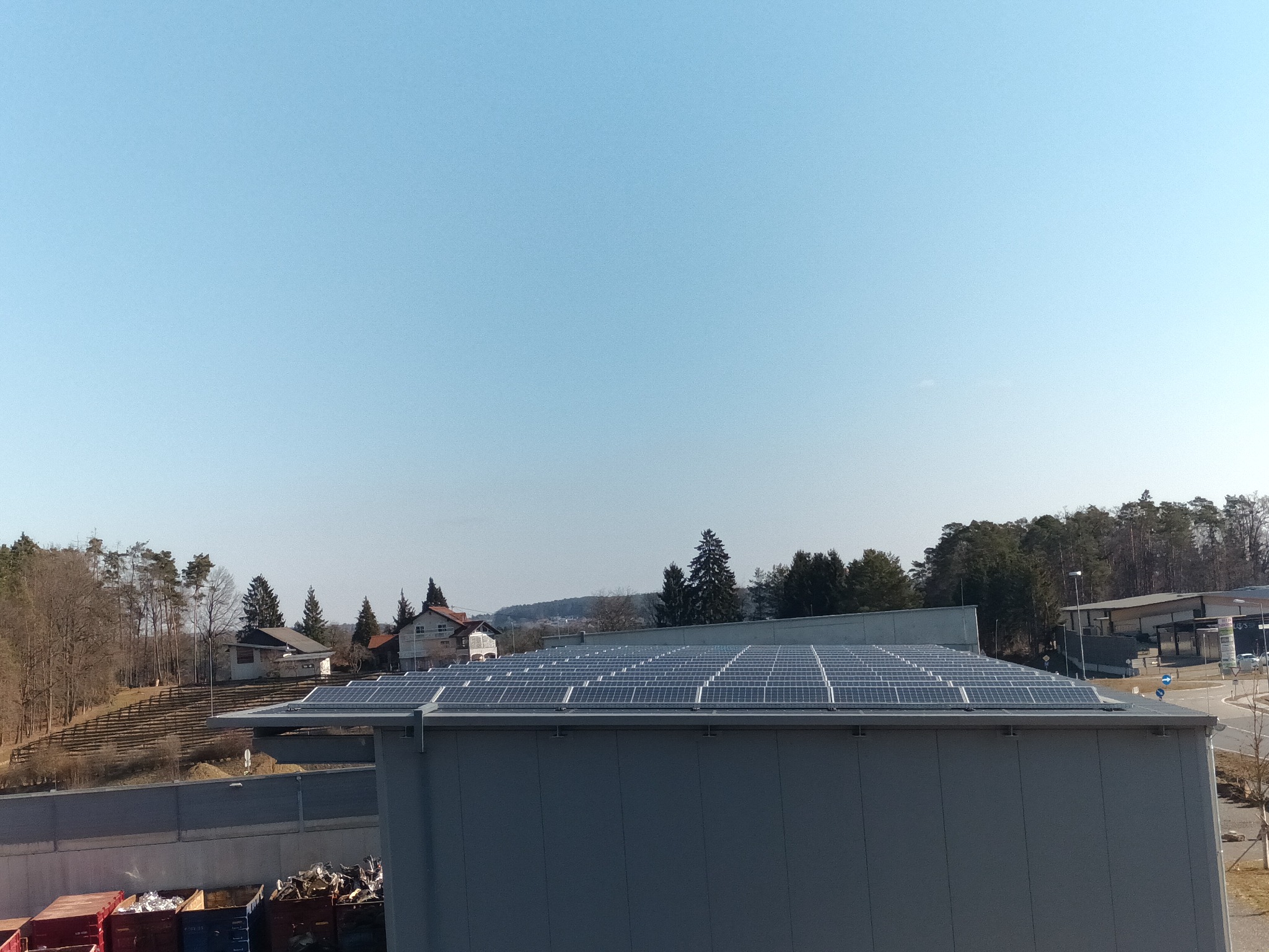 Równomiernie ułożone panele słoneczne na dachu budynku przemysłowego w słoneczny dzień, widok z góry na tle drzew i zabudowań.