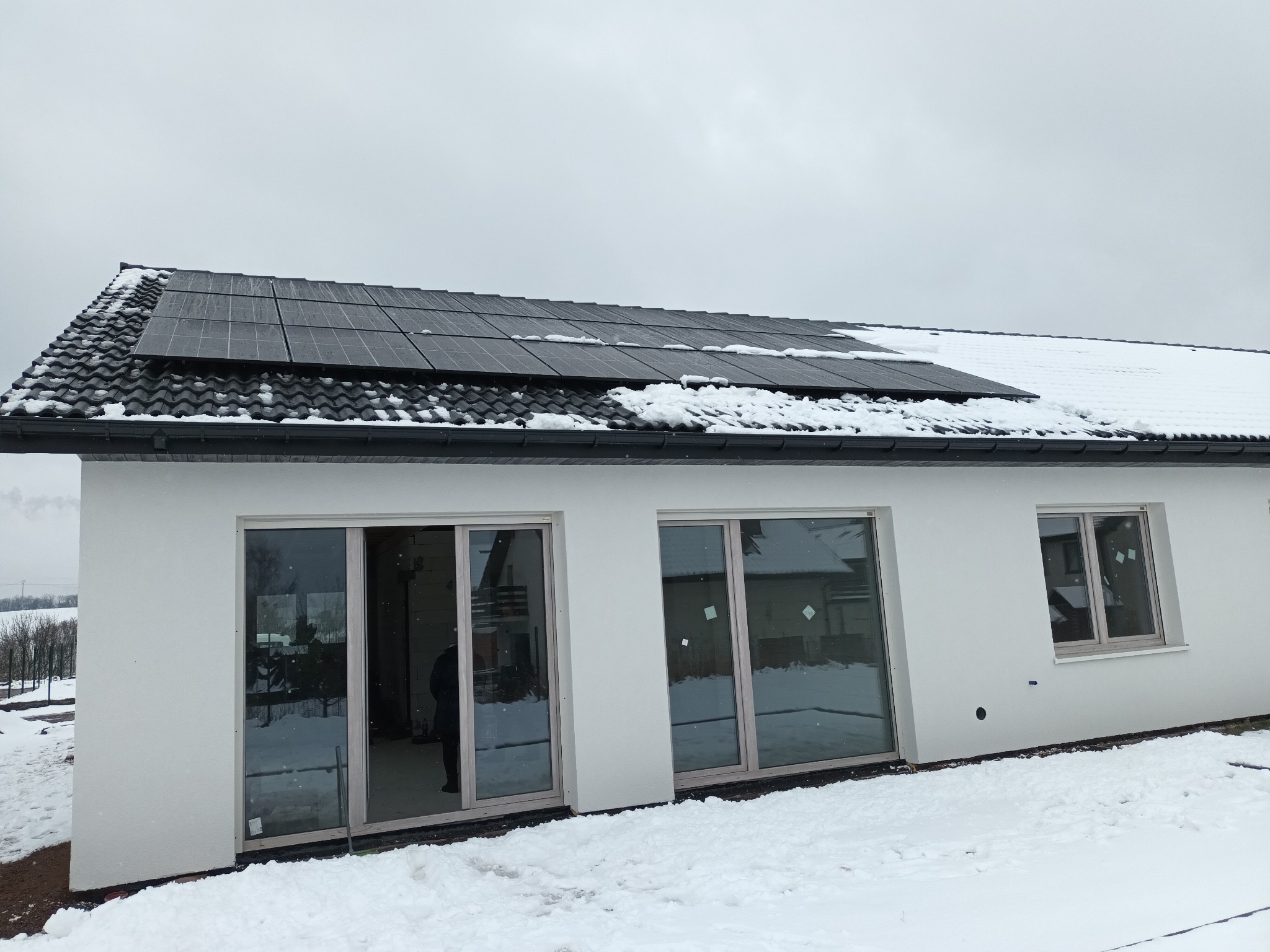 Biały dom z panelami słonecznymi na dachu pokrytym śniegiem, widok z zewnątrz w pochmurny dzień.