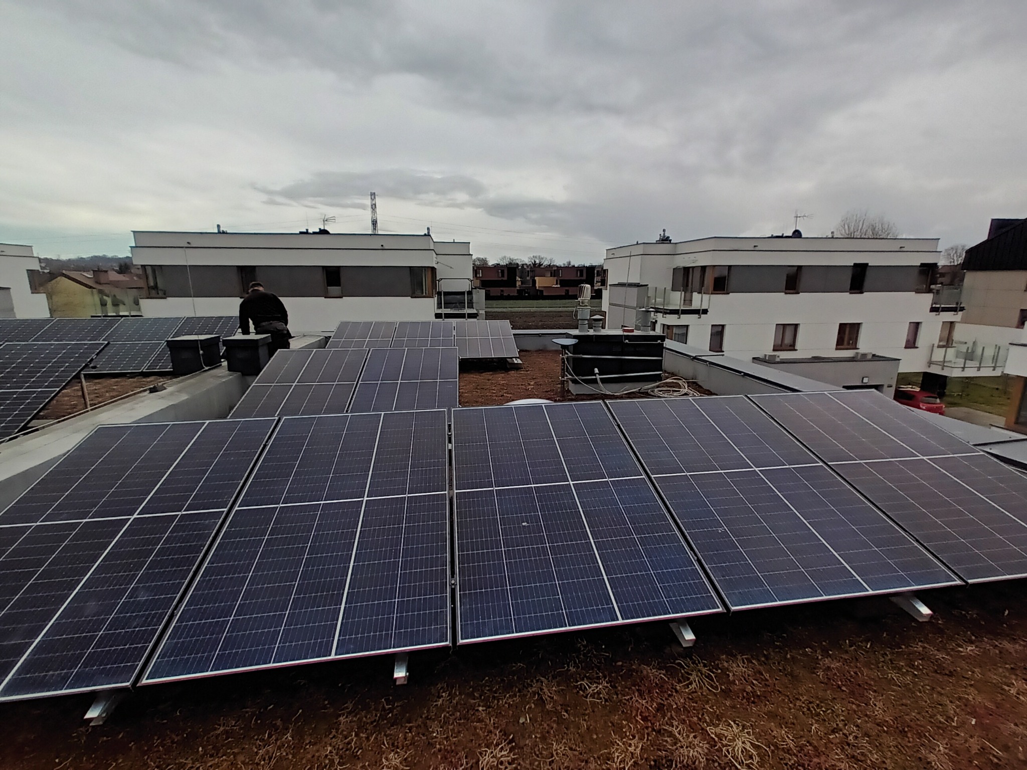 Instalacja paneli słonecznych na płaskim dachu budynku mieszkalnego, widoczny monter w ciemnej odzieży oraz sąsiednie budynki w tle, pochmurne niebo.