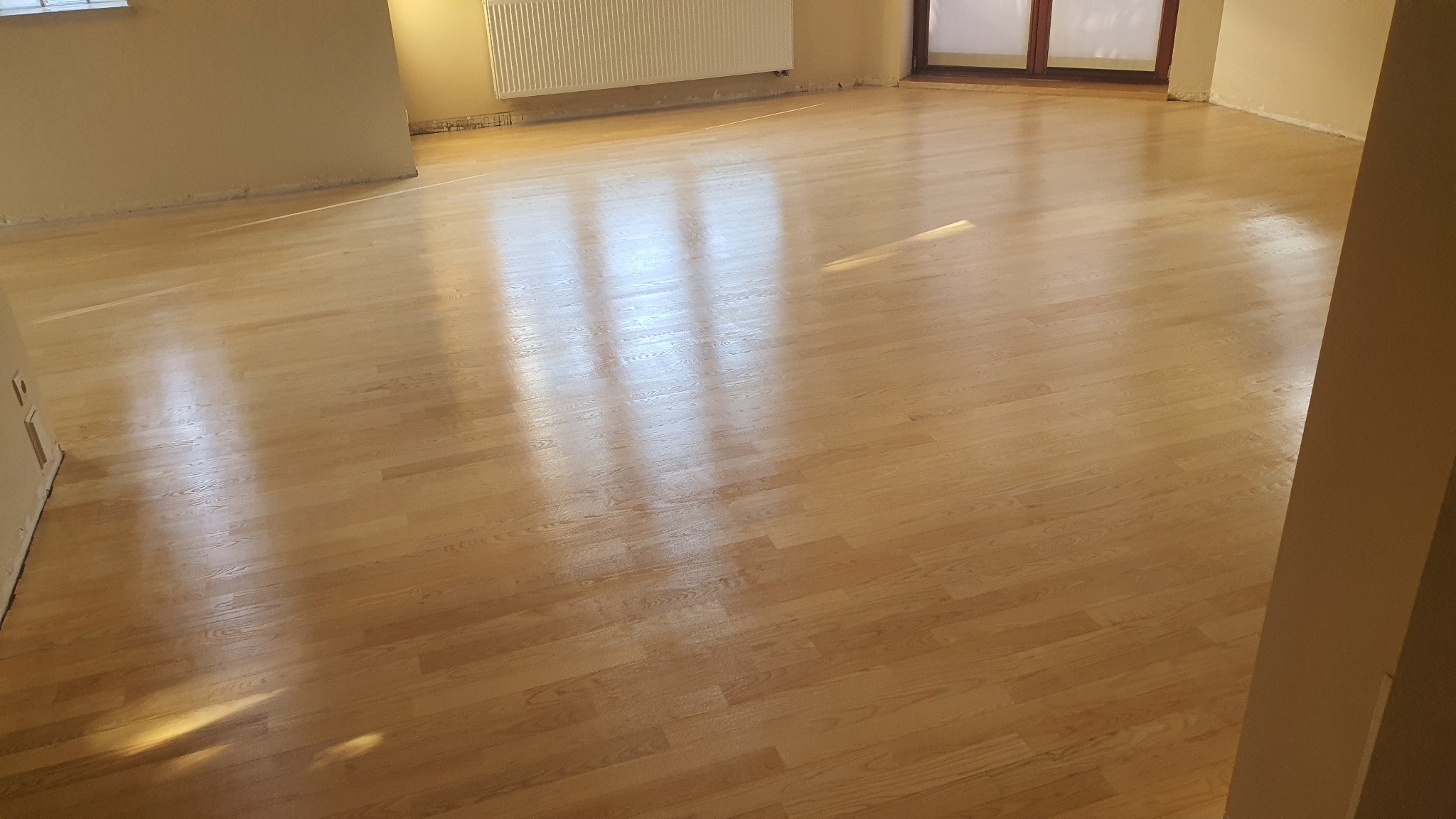 Wycyklinowana, jasna podłoga drewniana w pustym pokoju z widocznymi śladami po remoncie przy ścianach i grzejniku.