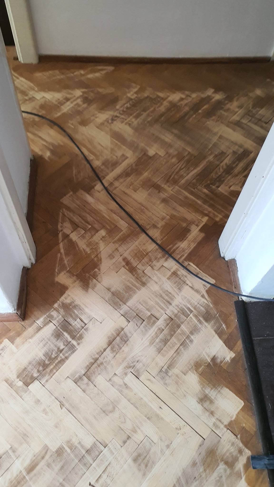 Parkiet w jodełkę częściowo wyszlifowany, widoczne różnice w odcieniach drewna, czarny kabel na podłodze, białe futryny drzwi