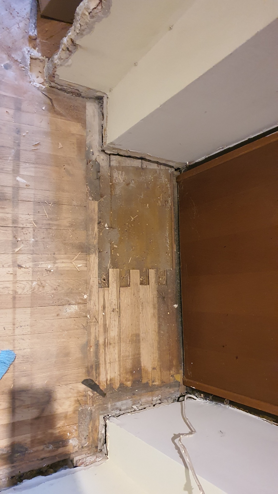 Fragment podłogi drewnianej w trakcie renowacji przy progu drzwiowym; widoczne stare deski, fragmenty uzupełnione nowym drewnem, ślady po kleju i ubytki w listwach przypodłogowych.