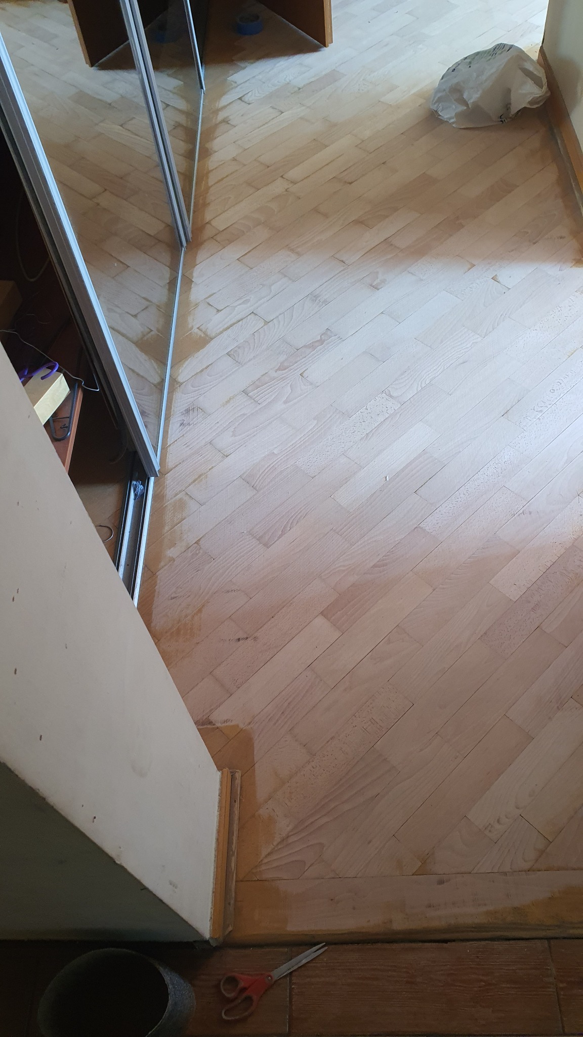 Parkiet ułożony w jodełkę po częściowym szlifowaniu, widoczne różnice w odcieniu drewna, przy listwie przypodłogowej leżą nożyczki i wiadro.