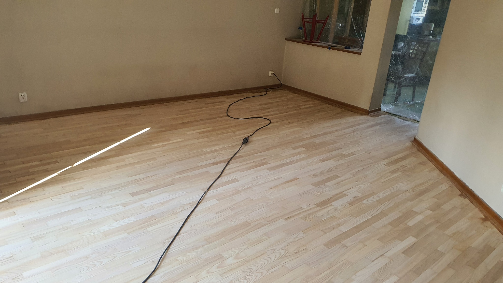 Jasna, odnowiona podłoga parkietowa w pustym pokoju przed malowaniem ścian, z widocznym kablem zasilającym na podłodze i oknem zabezpieczonym folią.