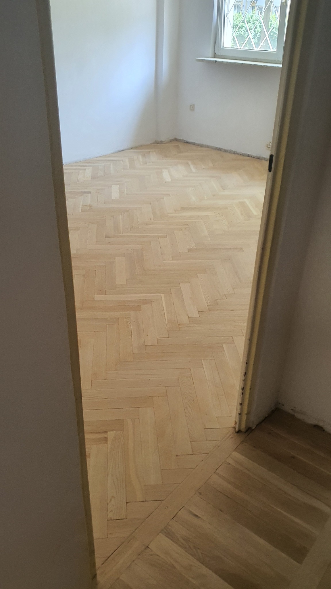 Jasna, odnowiona podłoga parkietowa ułożona we wzór jodełki w pustym pokoju, widoczna z perspektywy otwartych drzwi. Listwy przypodłogowe białe, ściany białe, okno z widokiem na zieleń w tle.