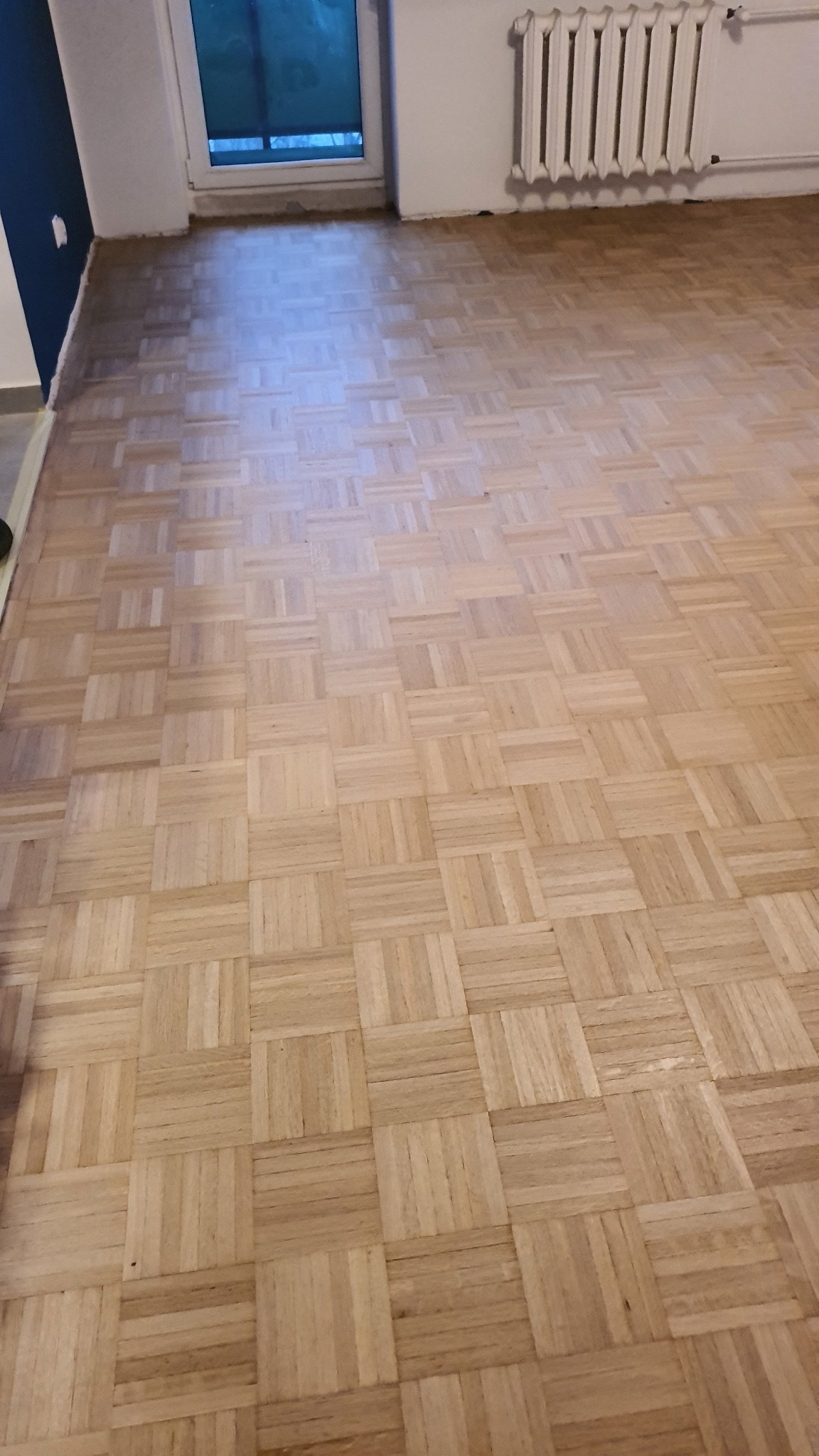 Parkiet ułożony w kwadraty po renowacji w mieszkaniu w Warszawie, widoczny grzejnik i fragment niebieskiej ściany.