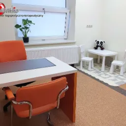 Gabinet logopedyczny z biurkiem, pomarańczowymi krzesłami, stolikiem dla dzieci z pluszową pandą i piankową podłogą z puzzli.