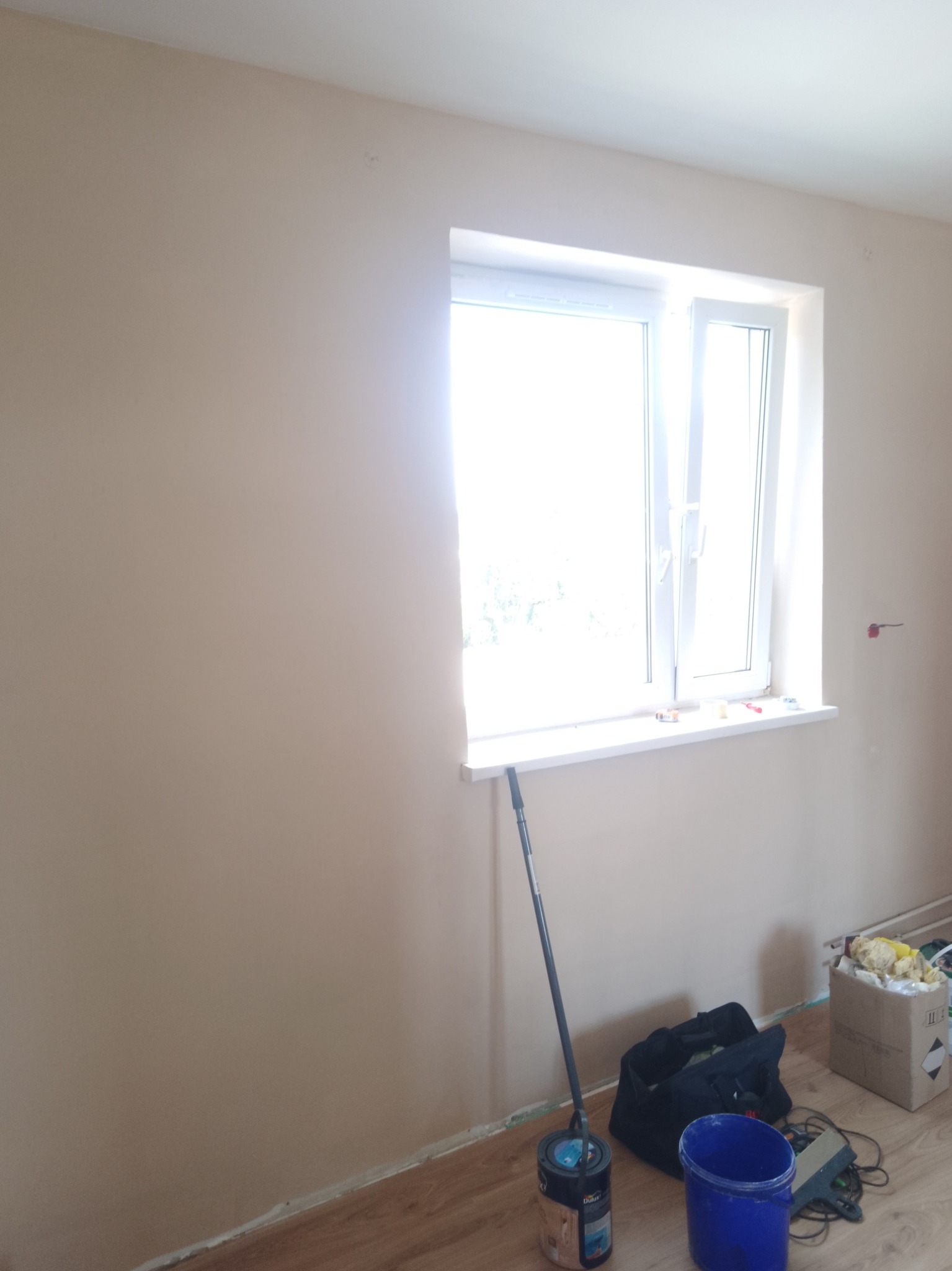 Świeżo pomalowane wnętrze pokoju z beżowymi ścianami, widoczne narzędzia malarskie: wałek oparty o ścianę, wiadro z farbą, niebieskie wiaderko, torba narzędziowa oraz karton z materiałami...