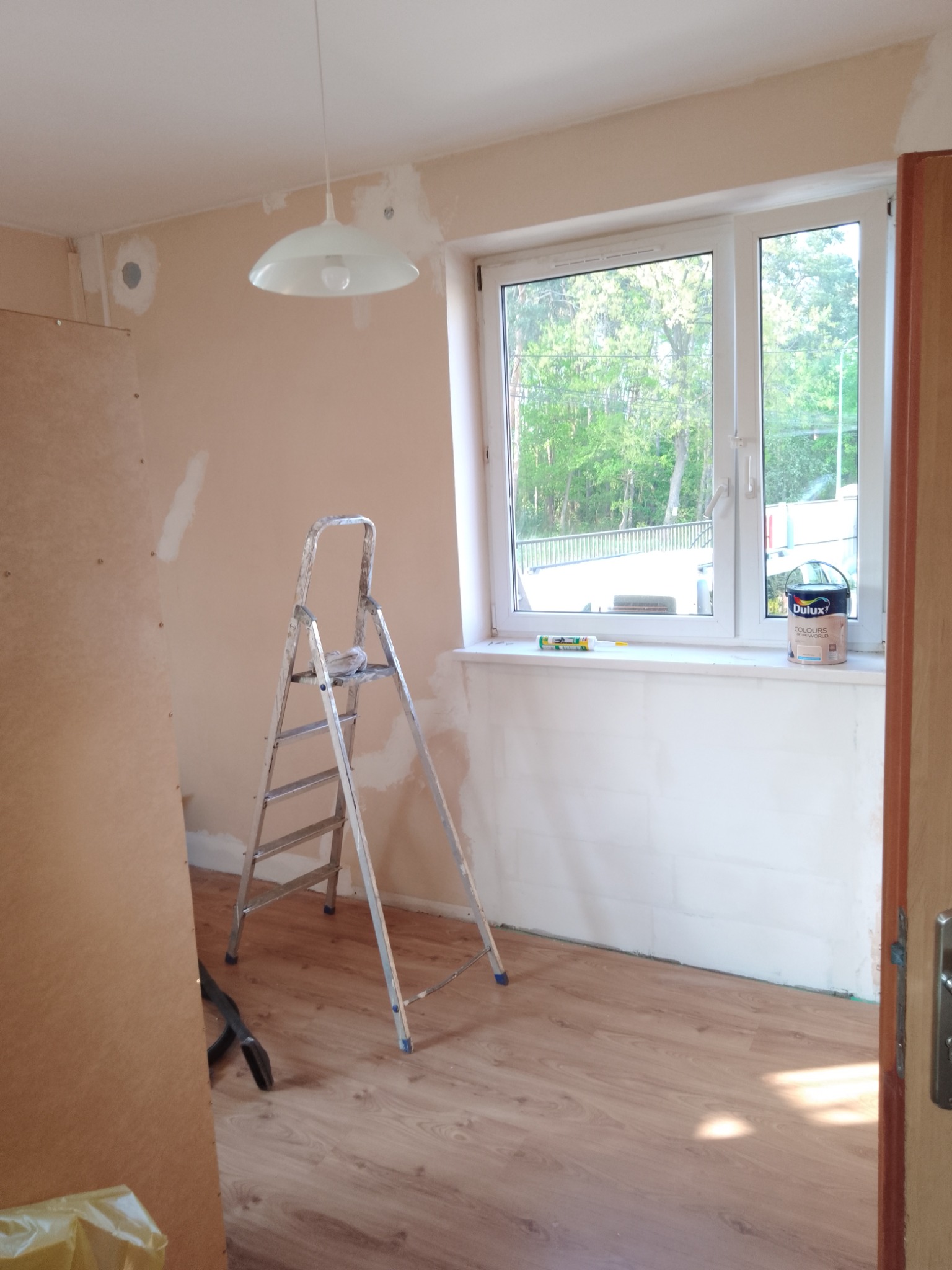 Pokój w trakcie przygotowania do malowania: ściany z fragmentami szpachli, aluminiowa drabina, puszka farby Dulux na parapecie, podłoga wyłożona panelami.