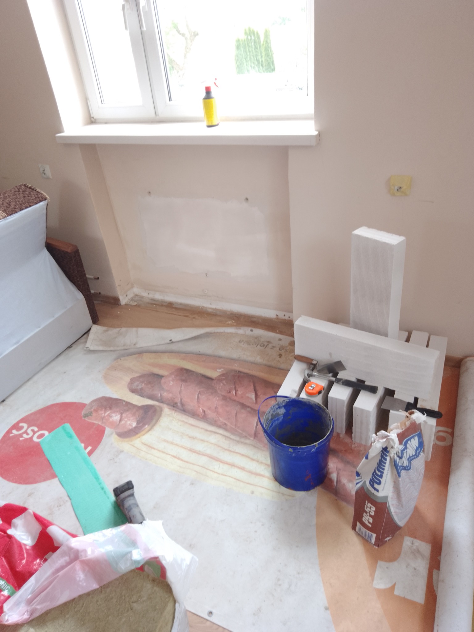 Przygotowania do ocieplania poddasza: styropian, wiadro z zaprawą, paca, poziomica i rolka folii ochronnej na podłodze w pobliżu okna.