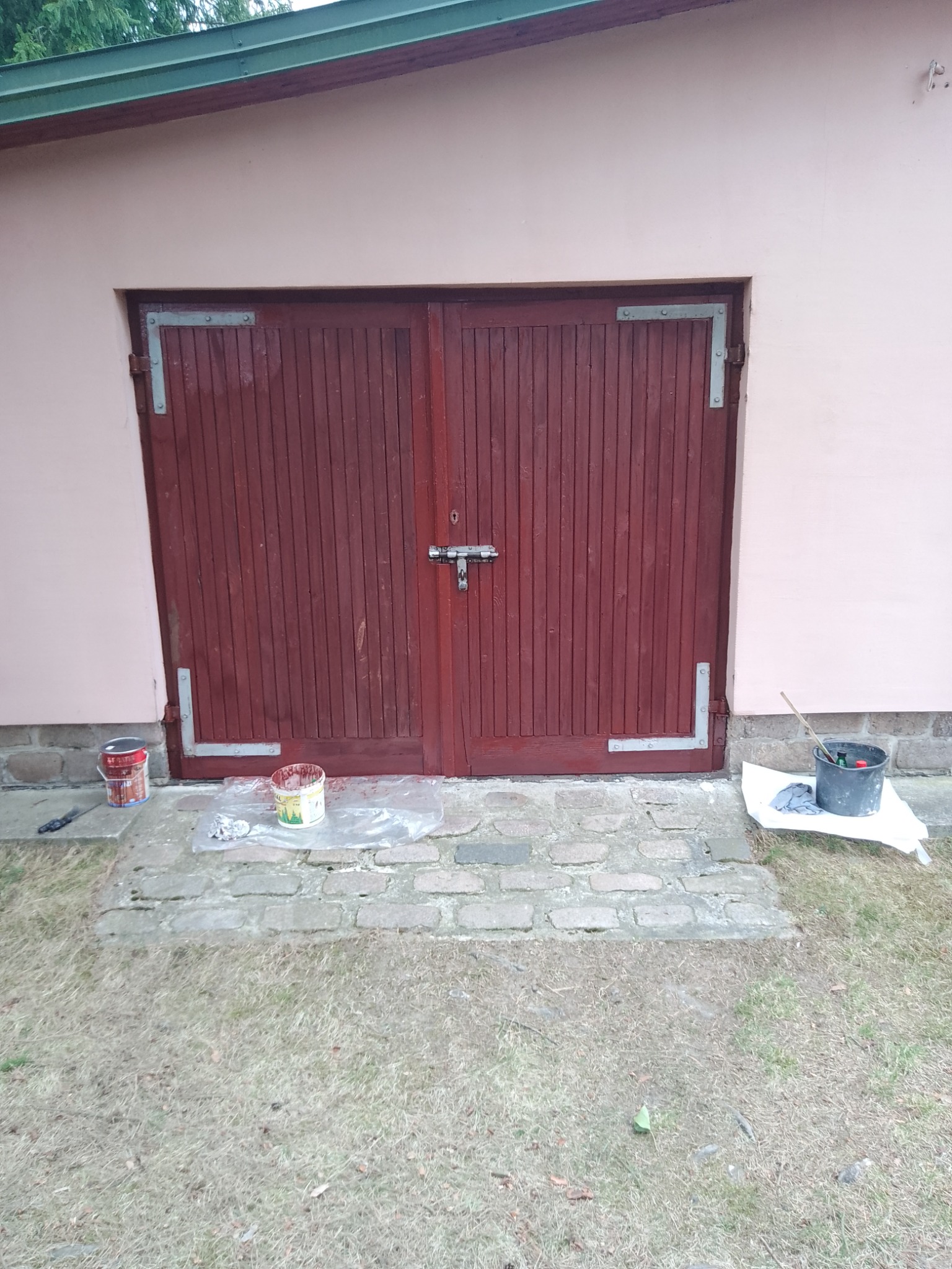 Ciemnoczerwone, drewniane, dwuskrzydłowe drzwi garażowe z metalowymi okuciami, po renowacji, z widocznymi puszkami farby i akcesoriami malarskimi na zewnątrz.
