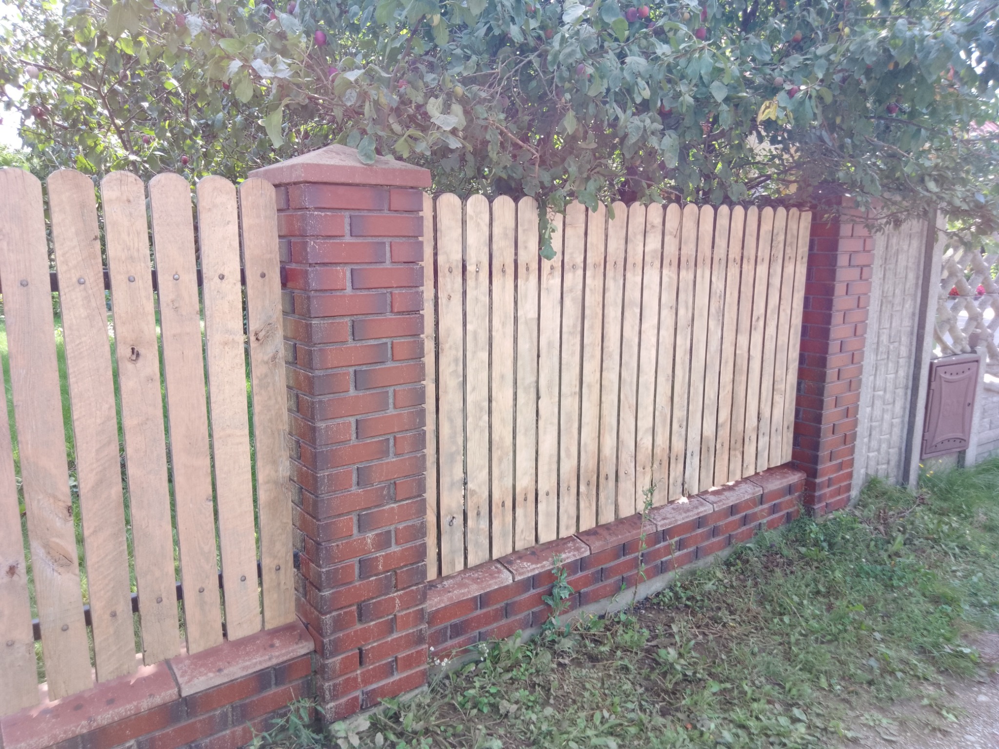 Nowy drewniany płot z ceglanymi słupkami i podmurówką, widoczny fragment drzewa z owocami w tle.