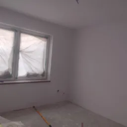 Przygotowane do malowania pomieszczenie w Radomsku, okna zabezpieczone folią, widoczny wałek malarski i wiadro farby.