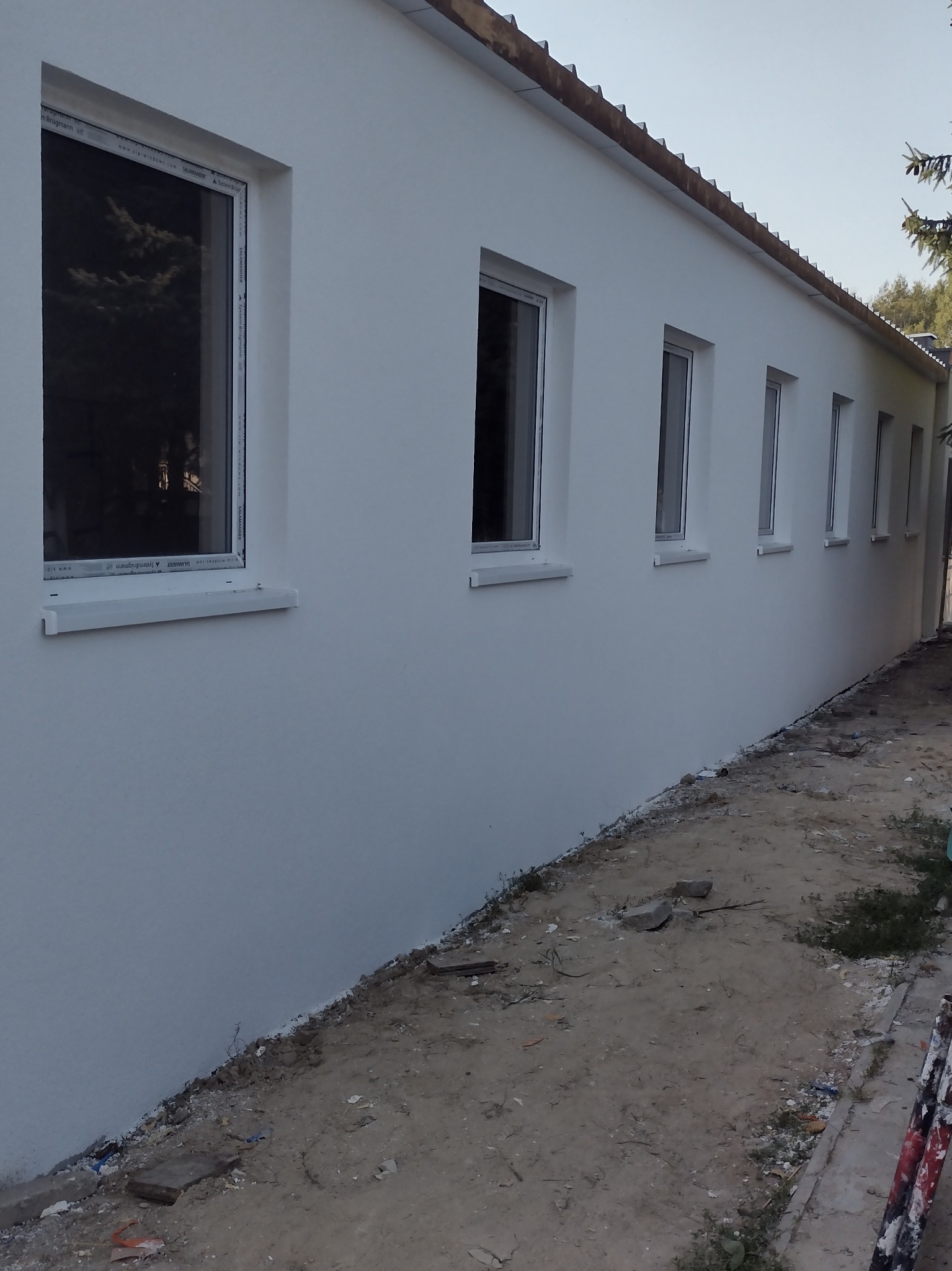 Biała elewacja budynku z rzędem okien z białymi ramami, widoczna z boku, z nierównym terenem u podstawy ściany i fragmentem drzewa w tle po prawej stronie.