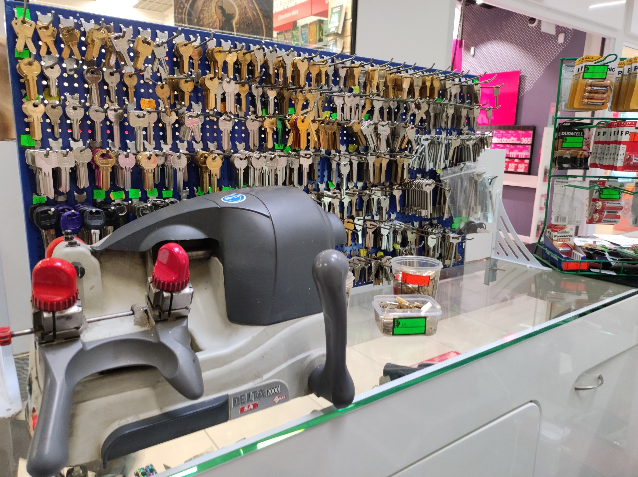 Maszyna do dorabiania kluczy Delta 2000 na tle perforowanej ściany z ekspozycją różnorodnych kluczy, widoczne pojemniki z kluczami do dorobienia.
