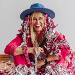 Uśmiechnięta blondynka w różowym garniturze i niebieskim kapeluszu, otoczona srebrno-różowym konfetti, trzyma mikrofon.