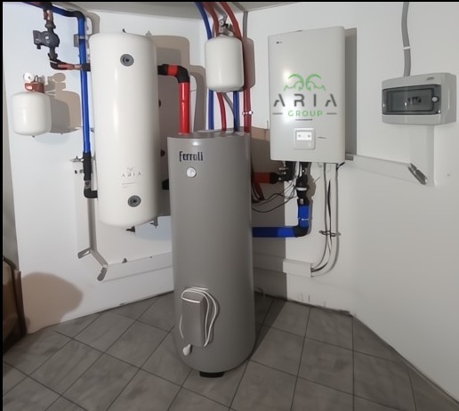 Kompletna instalacja grzewcza z szarym zasobnikiem Ferolli, białym zbiornikiem i jednostką sterującą Aria Group, z widocznymi rurami i skrzynką elektryczną.