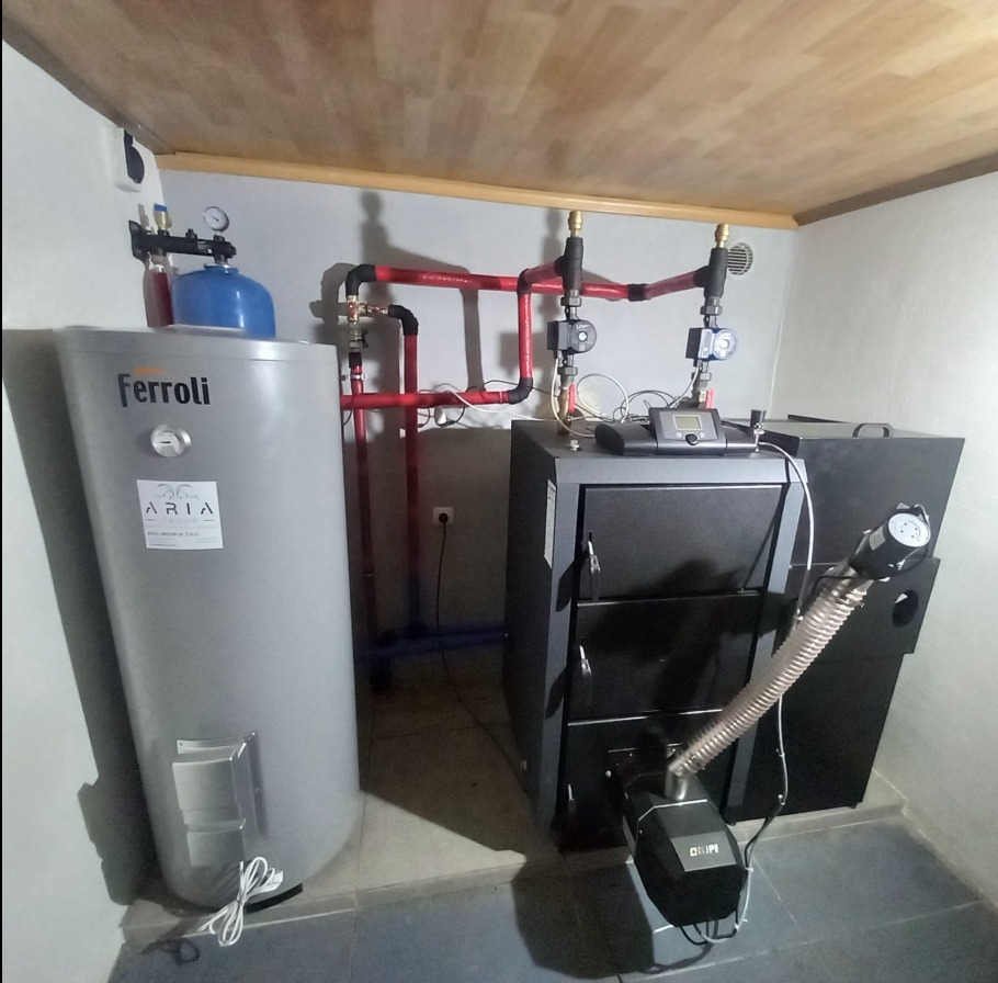 Zestaw urządzeń grzewczych w kotłowni: zbiornik Ferroli, piec na paliwo stałe z podajnikiem i instalacja z rur miedzianych. Widoczne elementy sterowania.