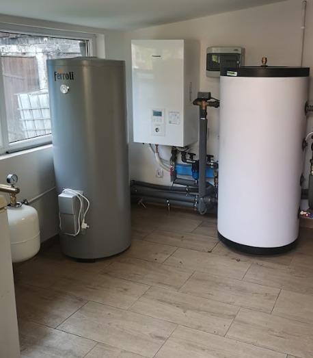 Zestaw urządzeń grzewczych w kotłowni: szary zasobnik Ferroli, biały piec Panasonic i biały zbiornik. Widoczne rury i armatura. Podłoga wyłożona płytkami imitującymi drewno.