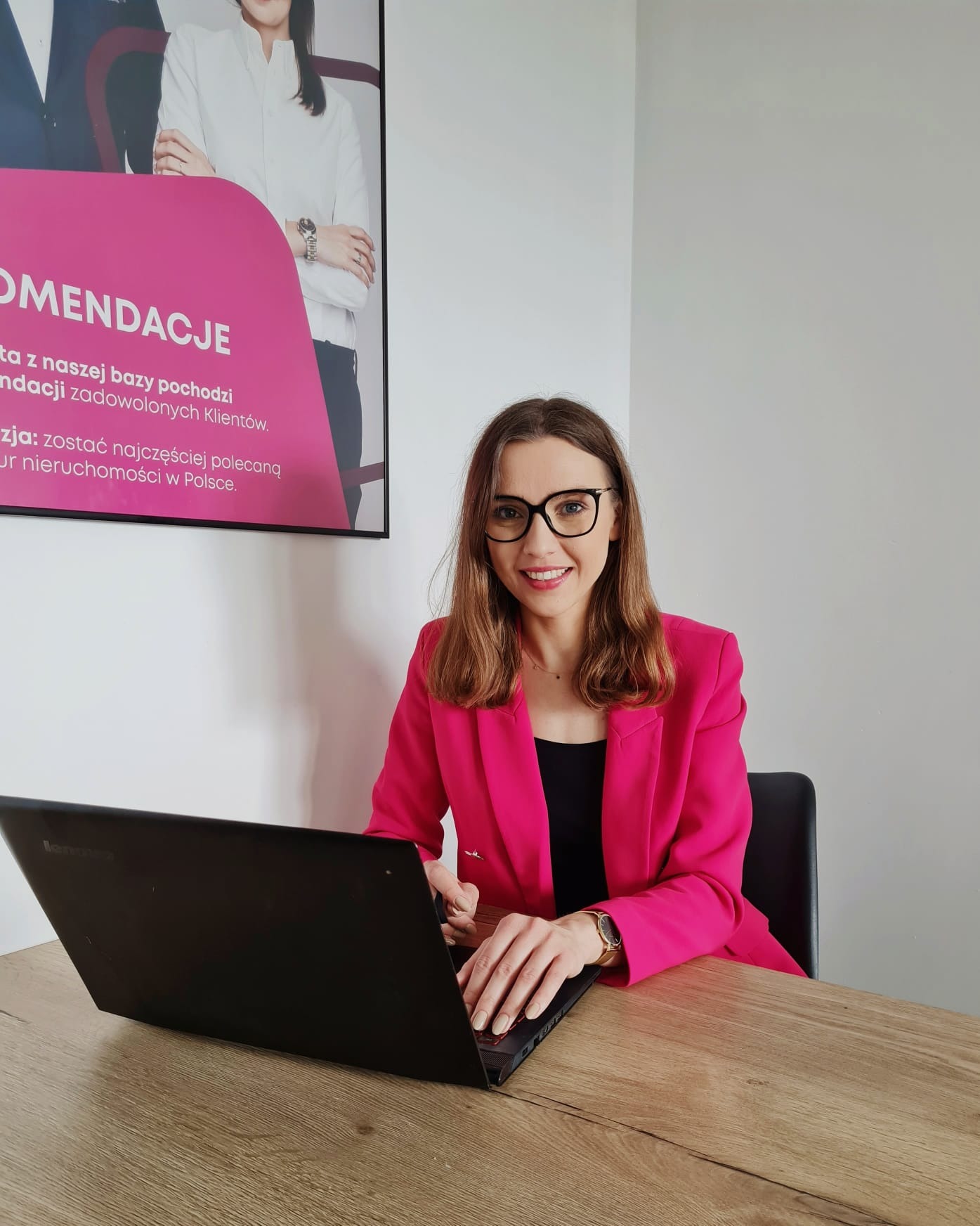 Uśmiechnięta agentka nieruchomości w różowej marynarce pracuje na laptopie przy biurku, w tle plakat z rekomendacjami firmy.