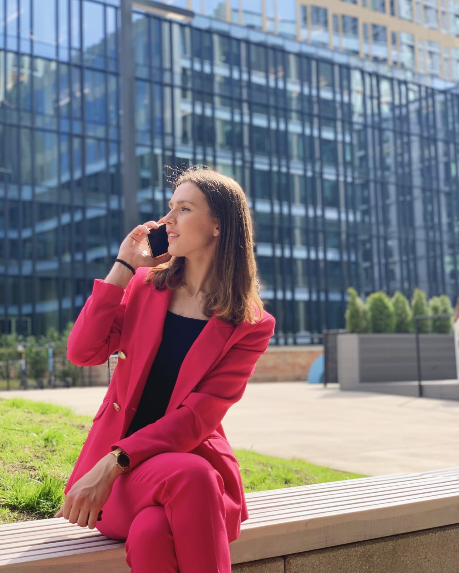Kobieta w różowym garniturze rozmawia przez telefon na ławce w Warszawie, na tle nowoczesnego biurowca.