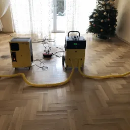 Dwa żółte osuszacze przemysłowe stoją na parkiecie ułożonym w jodełkę, jeden z widocznym panelem sterowania, drugi z dwoma elastycznymi wężami. W tle choinka i okno.