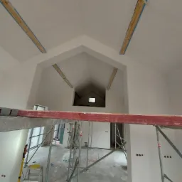 Wnętrze w trakcie wykańczania: białe ściany po tynkowaniu, widoczne rusztowania, drewniane elementy pod sufitem zabezpieczone niebieską taśmą malarską, przygotowane punkty elektryczne.