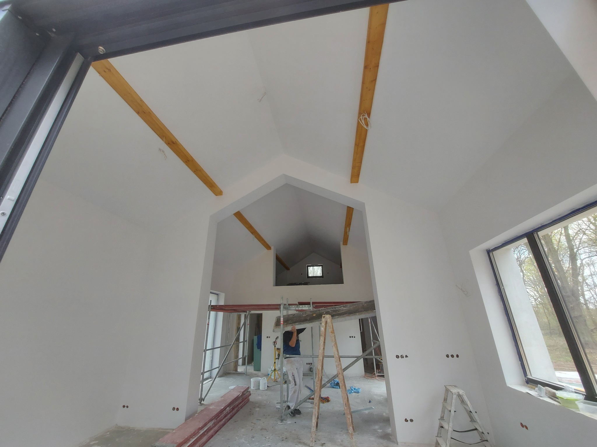 Wnętrze w trakcie prac wykończeniowych: świeżo otynkowane białe ściany i sufit, widoczne drewniane belki stropowe, rusztowanie i drabina, pracownik w oddali.