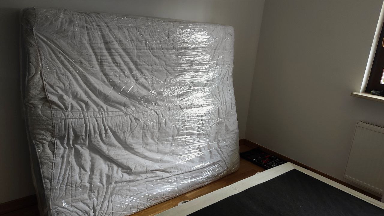 Materac owinięty folią ochronną, oparty o ścianę w rogu pokoju, przygotowany do transportu. Widoczny fragment ramy łóżka i grzejnika.