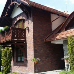 Dwukondygnacyjny budynek z czerwonej cegły z drewnianym balkonem i dekoracyjnymi elementami architektonicznymi, otoczony zielenią.