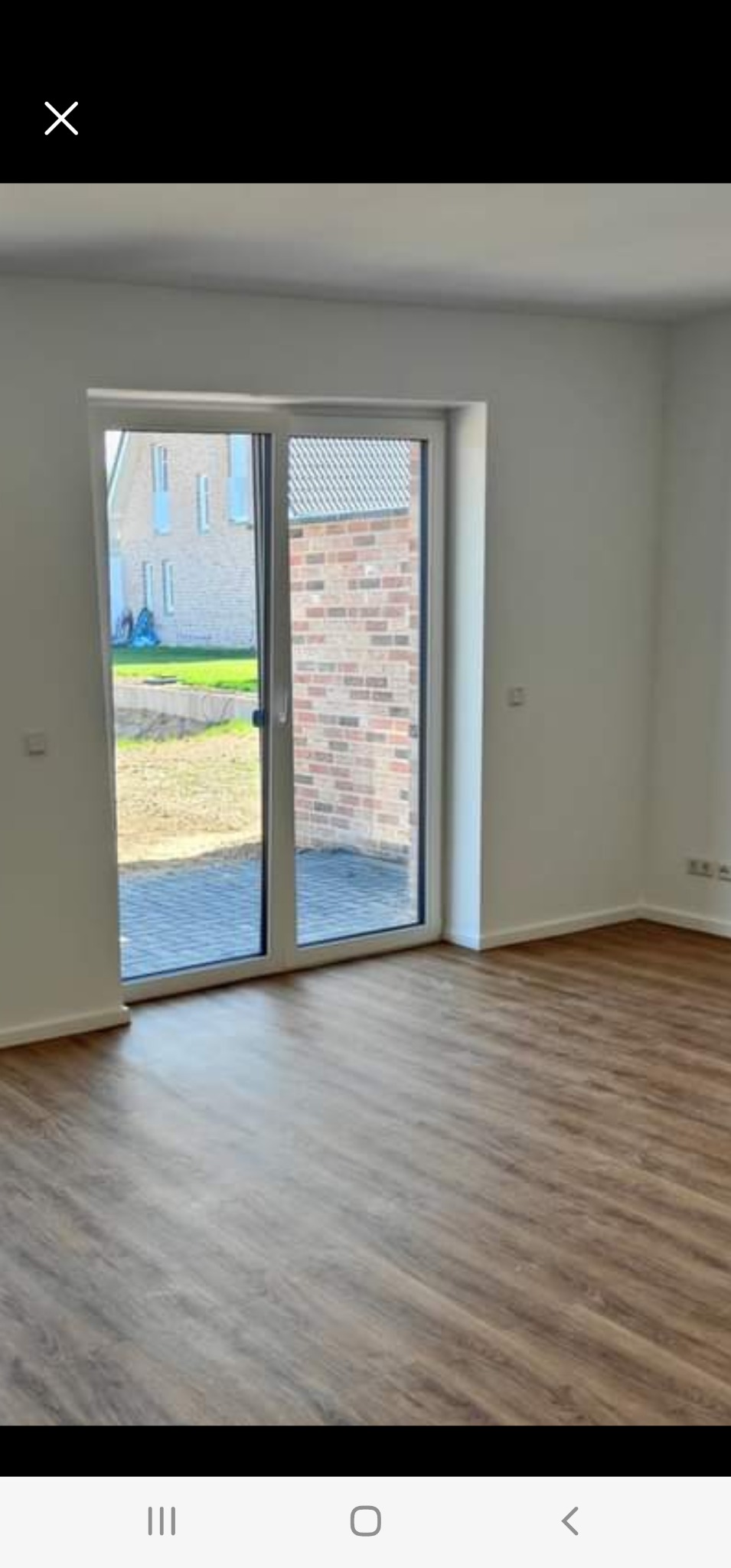 Wnętrze pokoju z panelami podłogowymi i przesuwnymi drzwiami balkonowymi z widokiem na ogród i budynek z cegły.