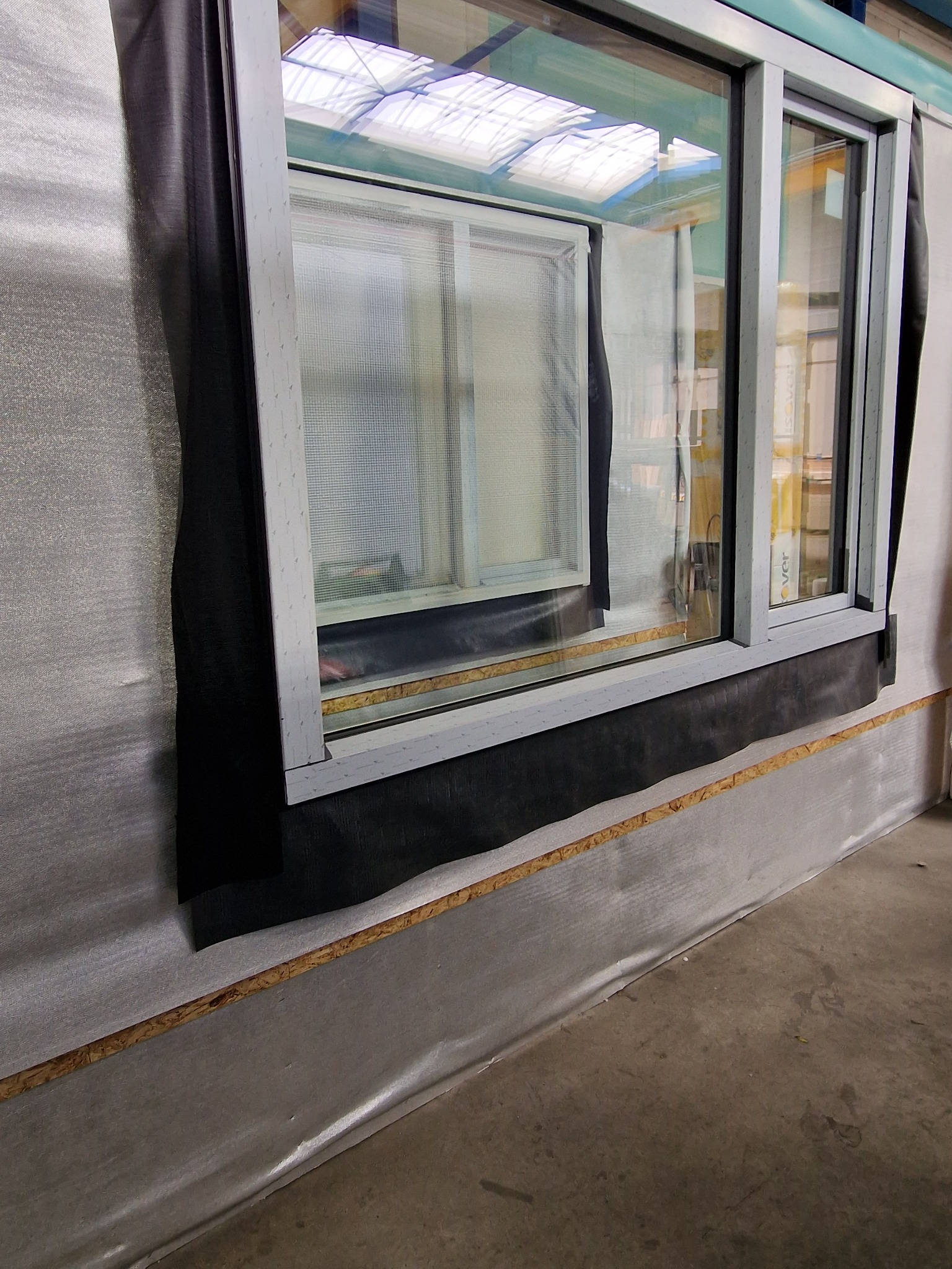 Montaż okna z szarą ramą, widoczne zabezpieczenia folią i taśmą uszczelniającą, częściowo zasłonięte czarną folią ochronną.