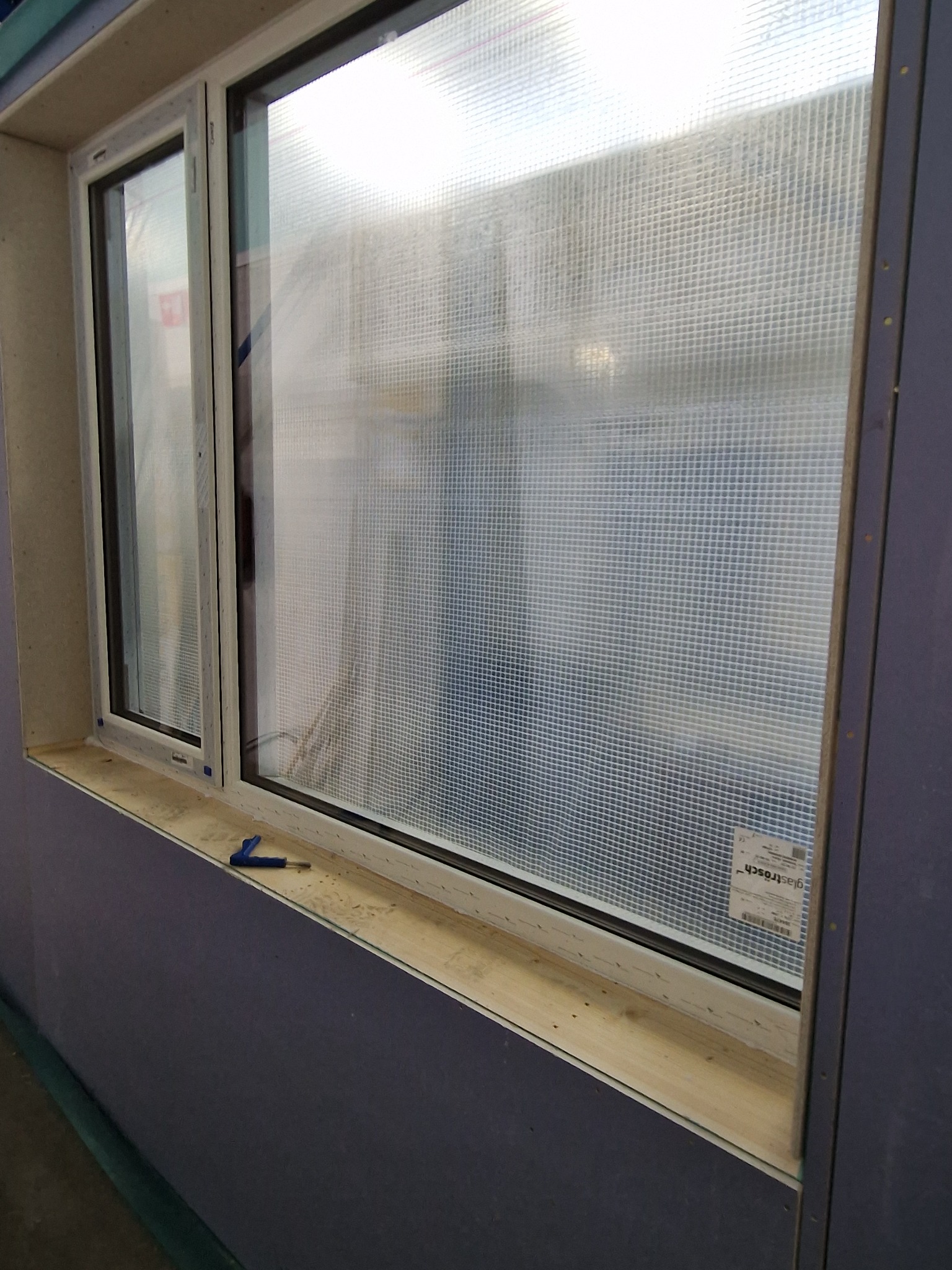 Nowe okno z szybą zabezpieczoną folią ochronną, widoczne narzędzie montażowe na parapecie, ściana w trakcie wykończenia.