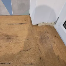 Fragment blatu drewnopodobnego, połączonego z płytkami w geometryczne wzory i białą ścianą z widocznymi śladami prac wykończeniowych.