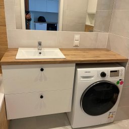 DREAM INTERIOR Elżbieta Chrupek - Biała umywalka nablatowa z chromowaną baterią, osadzona na blacie z jasnego drewna, pod nią biała szafka z dwiema szufladami i czarnymi okrągłymi uchwytami, obok pralka Samsung, całość w łazience...