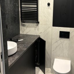 DREAM INTERIOR Elżbieta Chrupek - Czarna szafka łazienkowa z blatem imitującym kamień, biała umywalka nablatowa, wisząca toaleta i grzejnik drabinkowy na marmurowej ścianie.