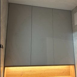 DREAM INTERIOR Elżbieta Chrupek - Szara szafka łazienkowa z trzema frontami, podświetlona od dołu ciepłym światłem, umieszczona na tle szarej ściany.