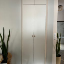 DREAM INTERIOR Elżbieta Chrupek - Wysoka, biała szafa z dwiema parami drzwi i okrągłymi, drewnianymi uchwytami, wkomponowana w korytarz, obok roślin doniczkowych i drewnianego krzesła. W tle widoczna łazienka z wanną i prysznicem.