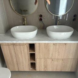 DREAM INTERIOR Elżbieta Chrupek - Podwójna umywalka nablatowa z marmurowym blatem i szafką z jasnego drewna, złote baterie i okrągłe lustra, widoczne niedokończone podłączenia elektryczne.