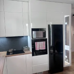 Nowoczesna kuchnia z białymi, lakierowanymi frontami, czarnym sprzętem AGD w zabudowie i drewnianym blatem, uzupełniona o płytę indukcyjną z garnkiem i koszyk z przyprawami.