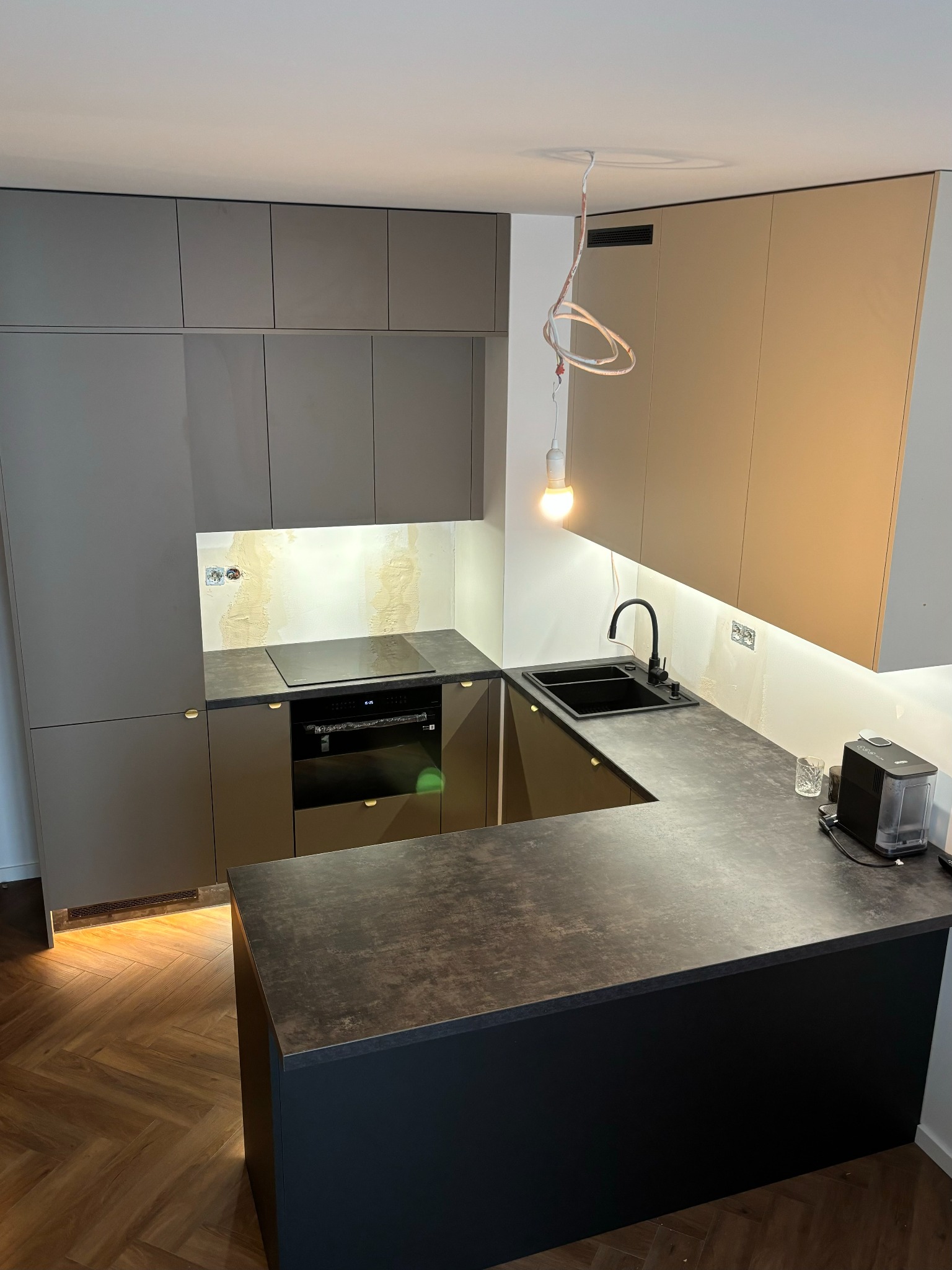 Nowoczesna kuchnia z szarymi i beżowymi szafkami, czarnym blatem i zlewozmywakiem, oświetleniem LED pod szafkami wiszącymi, oraz wiszącą żarówką na kablu, z widocznymi śladami prac wykończeniowych...